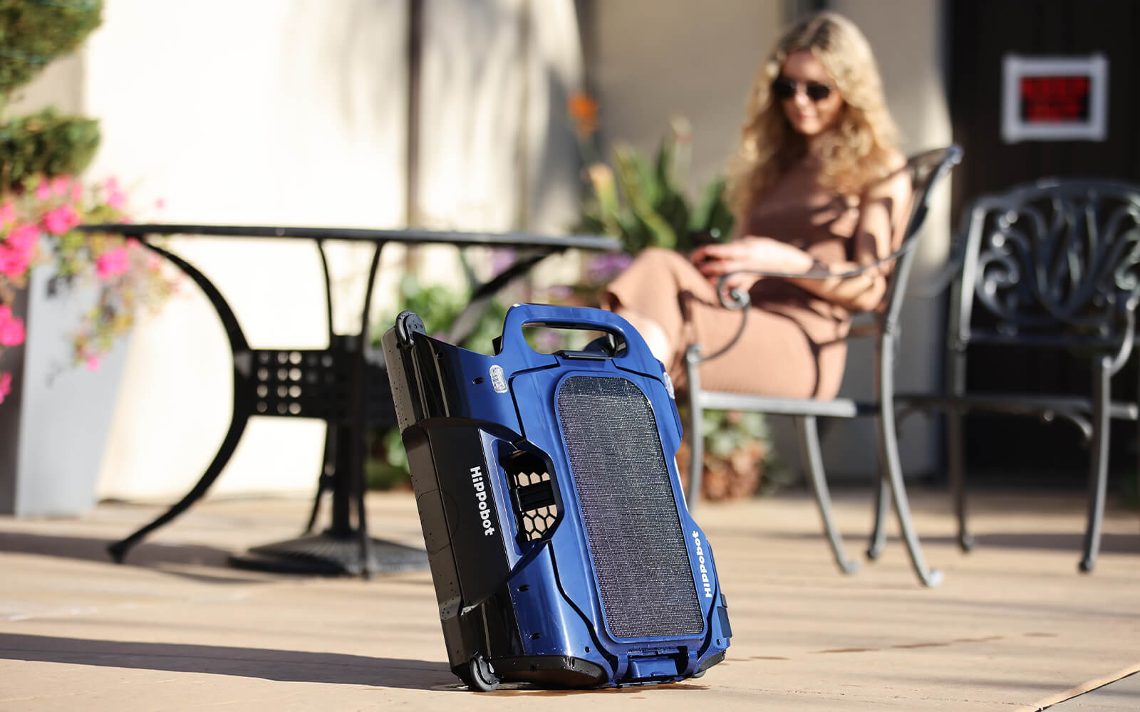 Hippobot H1 pool skimmer resting in a backyard, with a woman sitting beside the robotic cleaner.